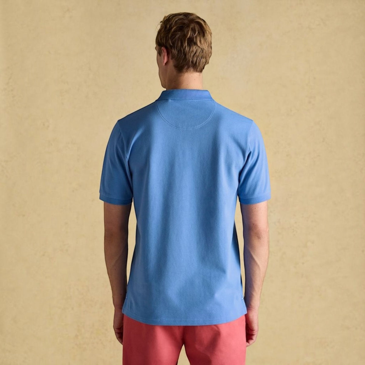 Back view of a man wearing a BATA LTD blue cotton pique polo shirt with short sleeves against a beige background.